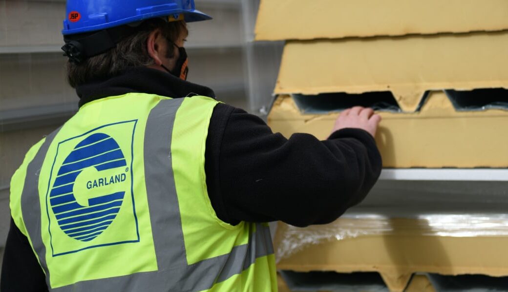 Worker wearing a high visibility vest with Garland logo and blue helmet moving a stack of yellow insulation boards