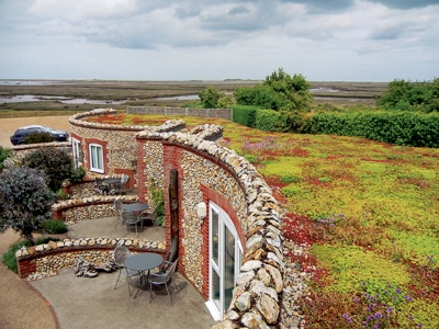 A curved stone house with a green, plant-covered roof overlooks a marshy landscape. Small patios with tables and chairs are outside each door, and a car is parked nearby. Dense greenery surrounds the area. Garland UK