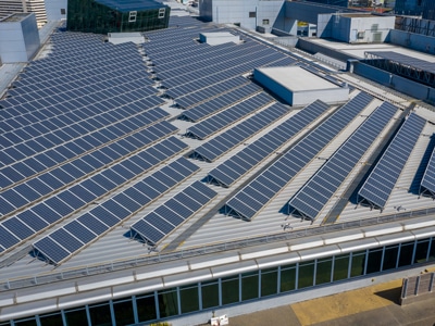 Rows of solar panels cover the standing seam metal roofing of a large industrial or commercial building, surrounded by other modern buildings in an urban setting, showcasing a commitment to renewable energy. Garland UK