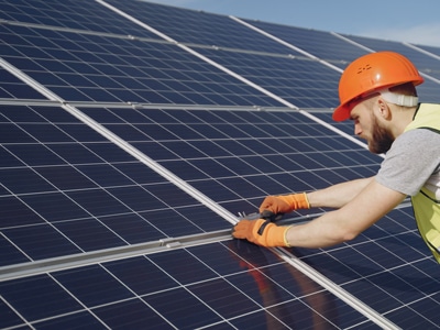 A worker wearing an orange helmet and gloves installs or inspects solar panels outdoors on a sunny day, gaining valuable Solar CPD experience. Garland UK