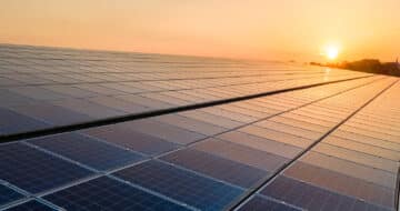 Rows of solar panels stretch across a roofing expanse, reflecting the warm light of a setting sun on the horizon, symbolizing renewable energy and sustainability. Garland UK