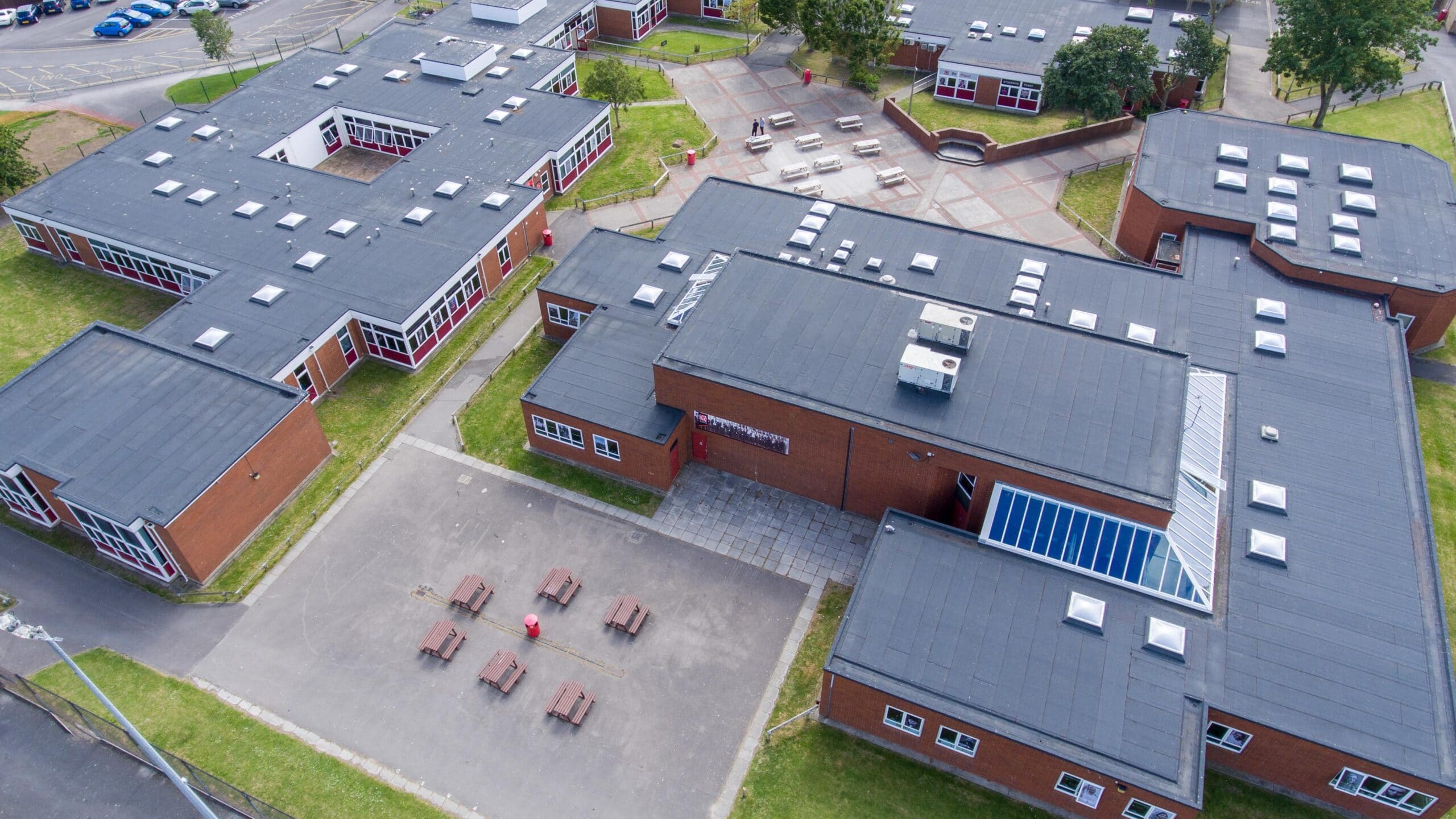 Aerial view of a school complex with multiple flat roofed red brick buildings, benches in a courtyard and surrounding trees
