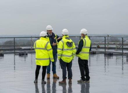Group of four construction workers wearing high visibility jackets and hard hats standing on a wet flat roof, one man gesturing as he speaks