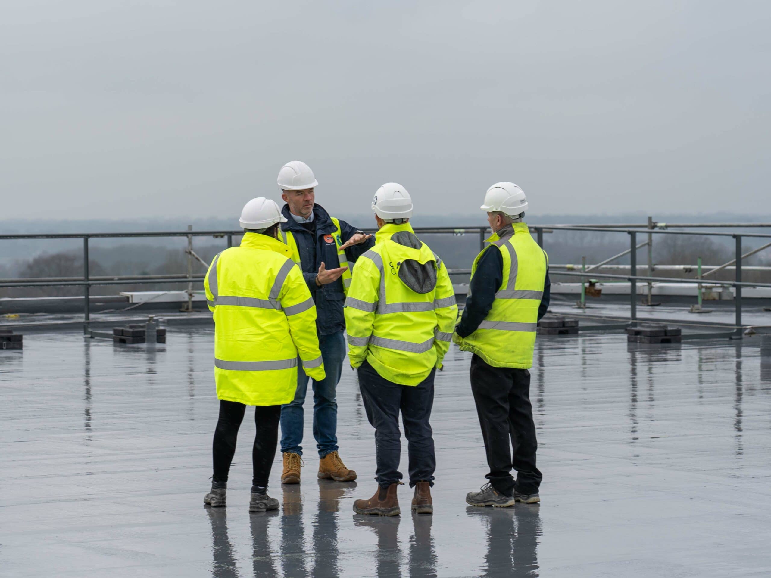 Group of four construction workers wearing high visibility jackets and hard hats standing on a wet flat roof, one man gesturing as he speaks