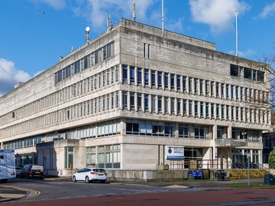 Exterior view of a multi storey concrete building with rows of rectangular windows and scaffolding along the ground floor, with cars parked and a blue sky above