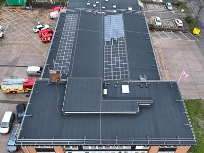 Aerial view of a flat roof with solar panels and vents covering much of the roof, surrounded by a brick building and car park