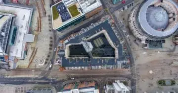 Aerial view of a city block showing roads, modern buildings with flat roofs, a circular building and a central courtyard, with traffic and pedestrians visible