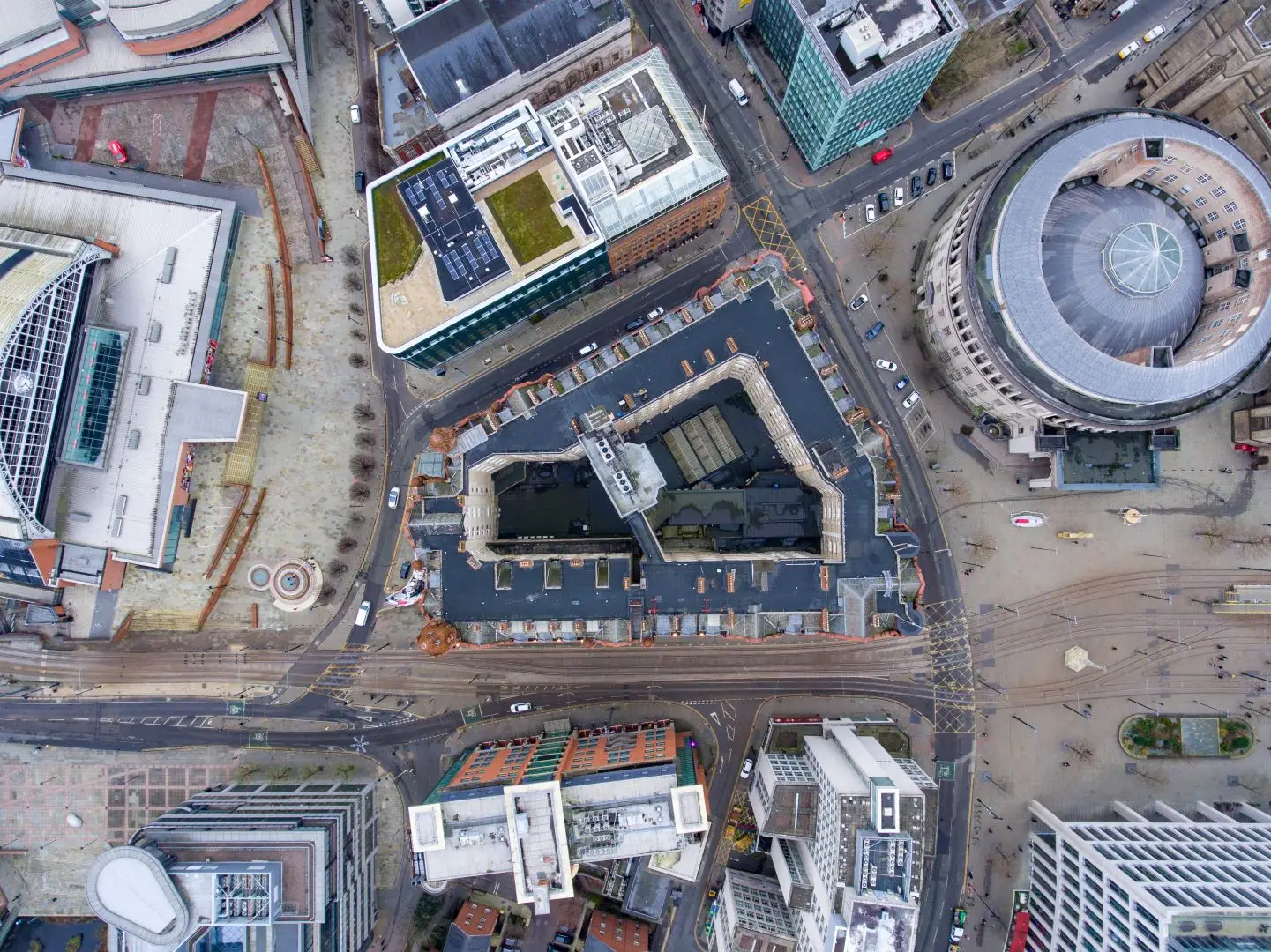 Aerial view of a city block showing roads, modern buildings with flat roofs, a circular building and a central courtyard, with traffic and pedestrians visible