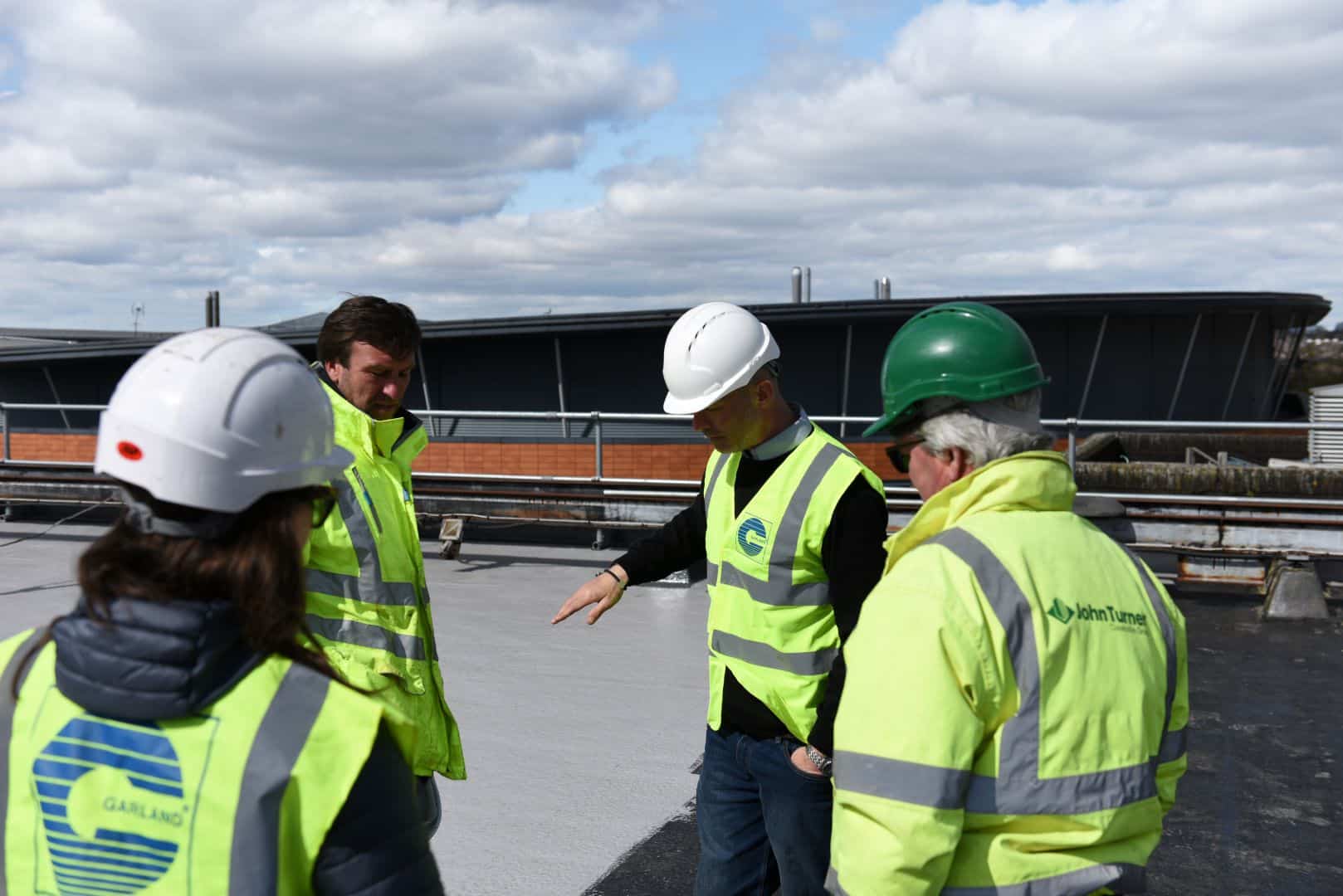 Group of four people wearing high visibility jackets and hard hats inspecting a flat roof, with a blue sky in the background