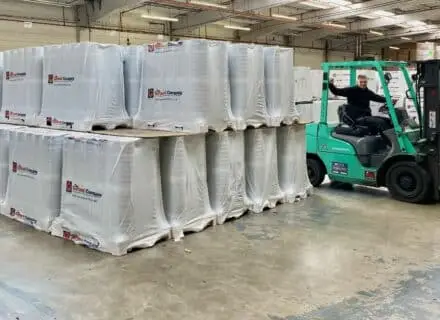Warehouse scene with stacks of wrapped pallets on the left and a man operating a teal forklift on the right inside an industrial building