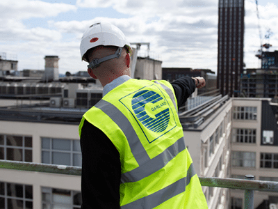 Rear view of a person wearing a white hard hat and high visibility vest with Garland logo pointing towards a building from a rooftop