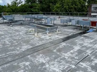 Flat commercial rooftop with aging, cracked waterproof membrane and various HVAC ducts and vents visible across the surface.