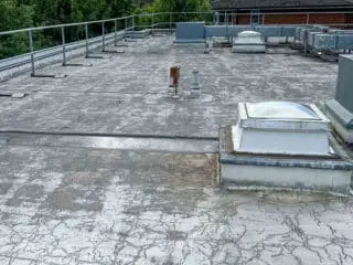 Worn flat roof showing significant surface cracking, with multiple skylights and rooftop equipment; surrounding guard rails line the perimeter.