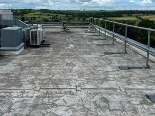 Weathered flat roof with peeling and deteriorated surface; air conditioning units and vents are positioned along the edge, overlooking green countryside.