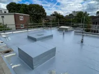 Rooftop mid-renovation with tools and buckets scattered around, surface partially stripped and being coated; a worker in high-visibility gear is present