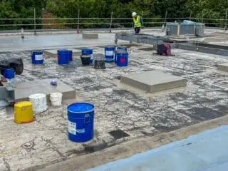 Close-up of a rooftop area being waterproofed; patches of coating contrast with exposed sections, and vent pipes protrude through the surface.