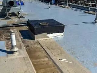 Partially refurbished roof with smooth waterproof membrane applied; ventilation unit sits on a raised section, with clear work-in-progress signs.